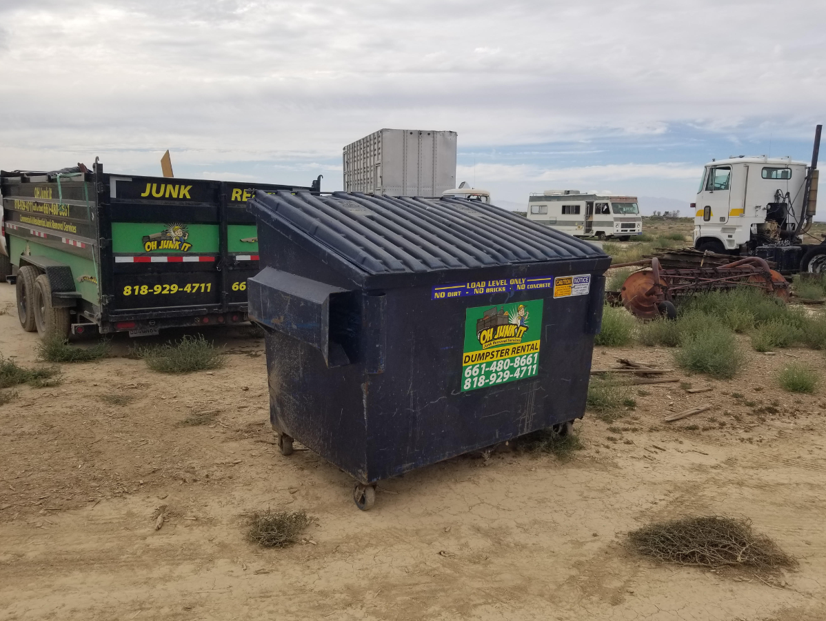 A dark blue dumpster with a junk hauling trailer, truck, and RVs in a field on a cloudy day.