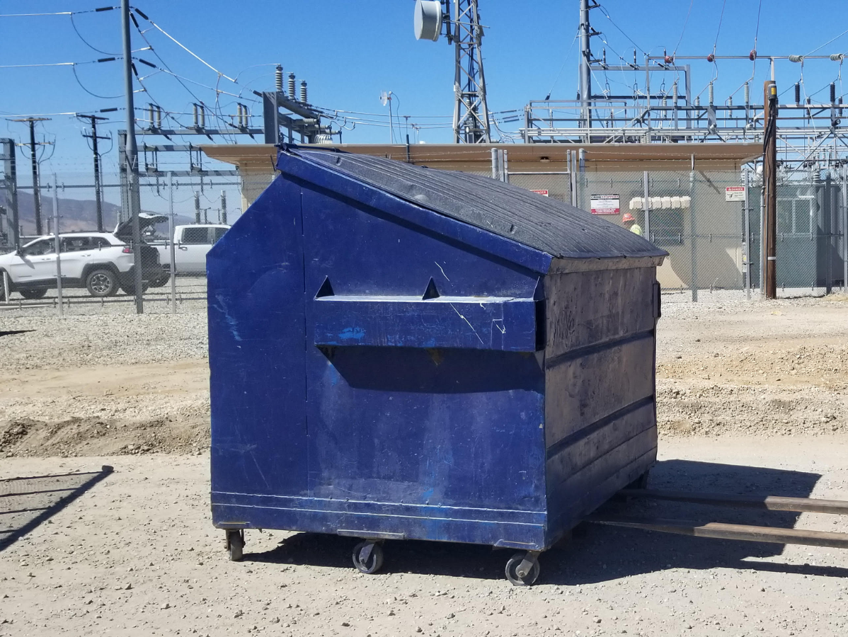 Blue dumpster on wheels near an industrial area, with power lines and vehicles in the background.