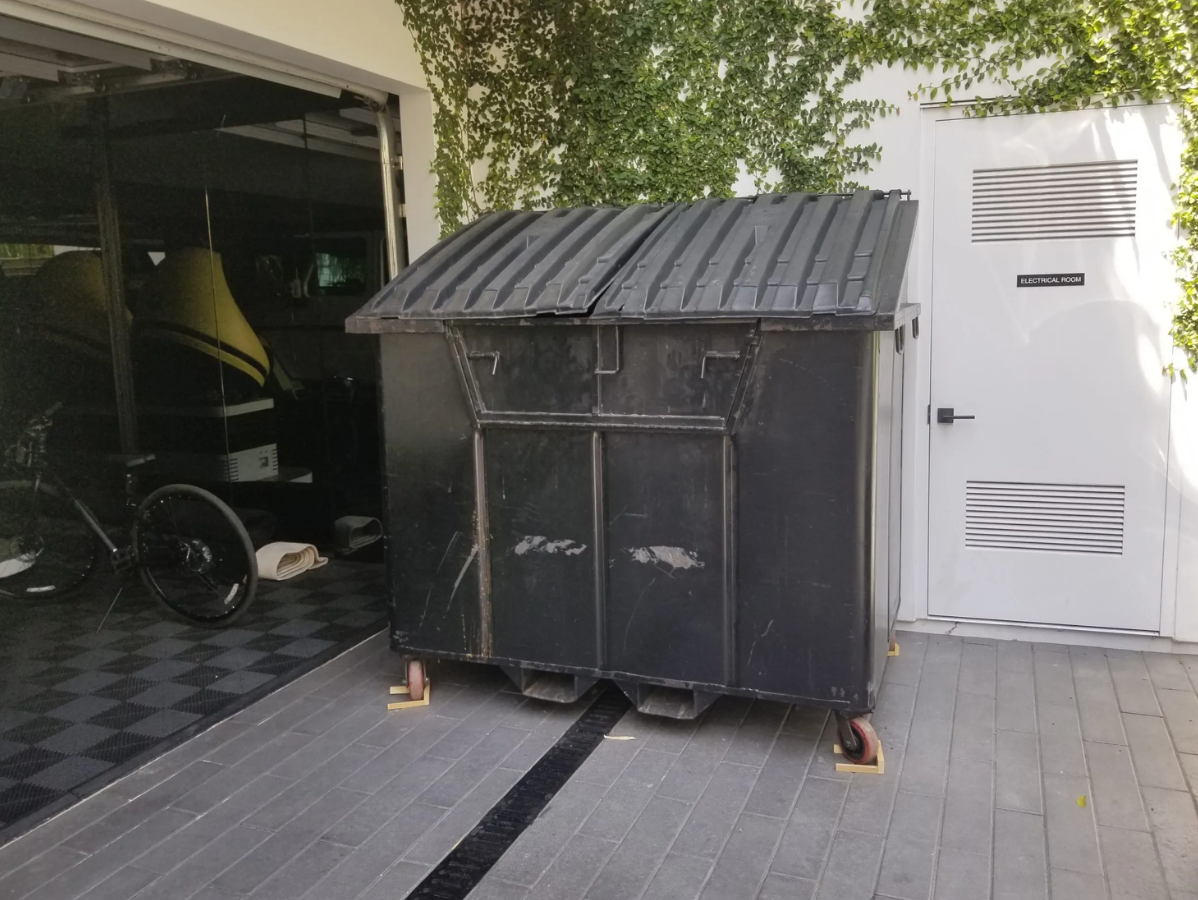 Black dumpster on wooden platform next to a white door and garage with bicycle.