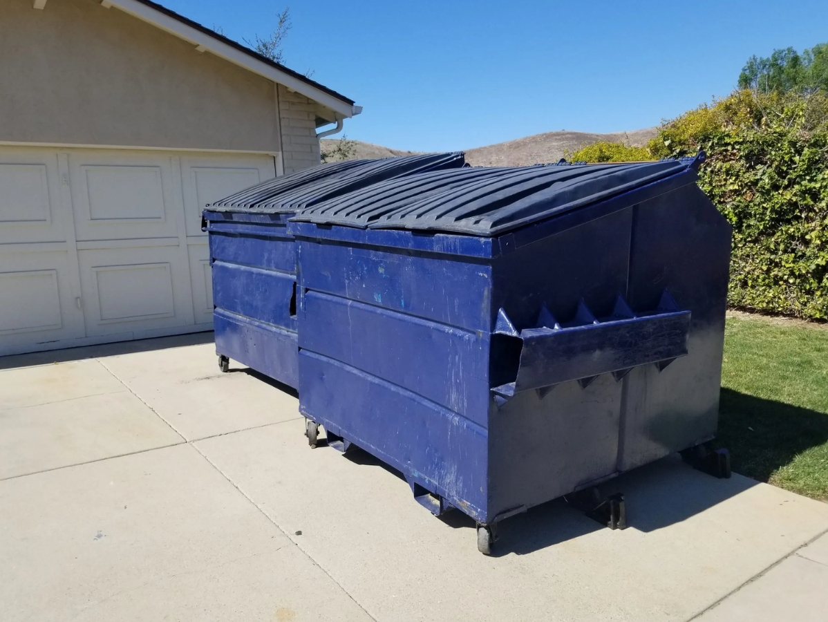 Blue dumpsters on wheels sit on a concrete driveway next to a house on a sunny day.