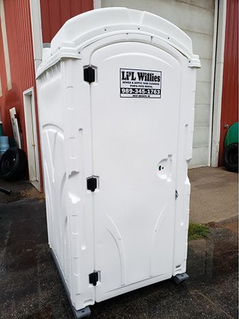 A white portable toilet is parked in front of a red building.