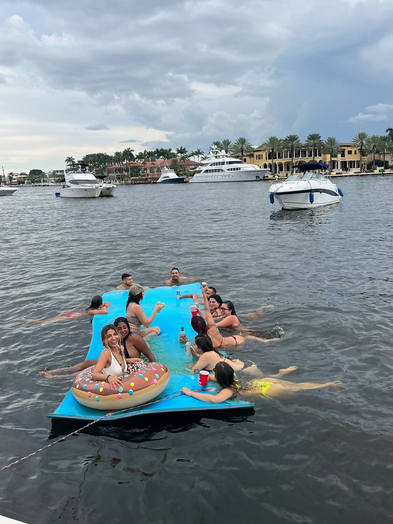 People on a blue floating raft in water, near boats, under a cloudy sky.