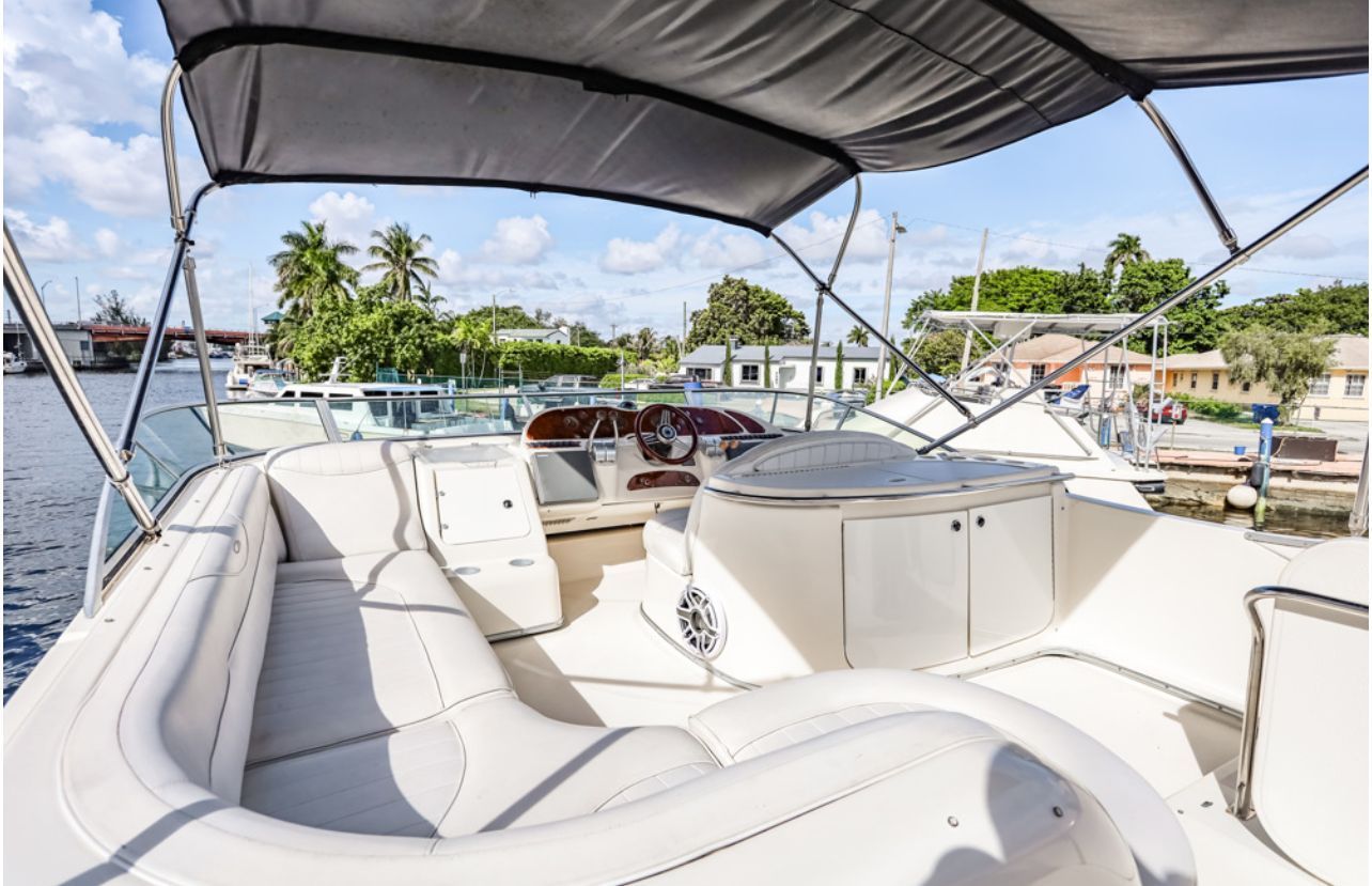 Boat interior with white seating and a steering wheel, docked on a sunny day with palm trees in the background.