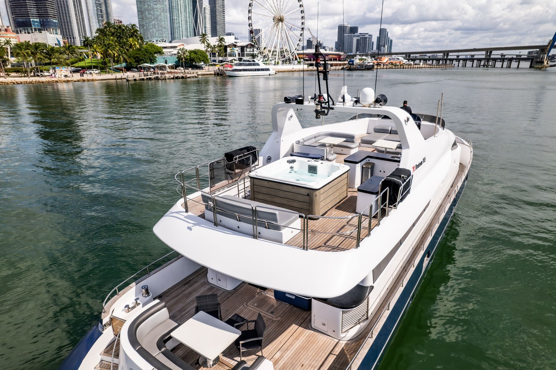 Luxury yacht with a jacuzzi on the deck, docked in a harbor with a Ferris wheel and city skyline in the background.