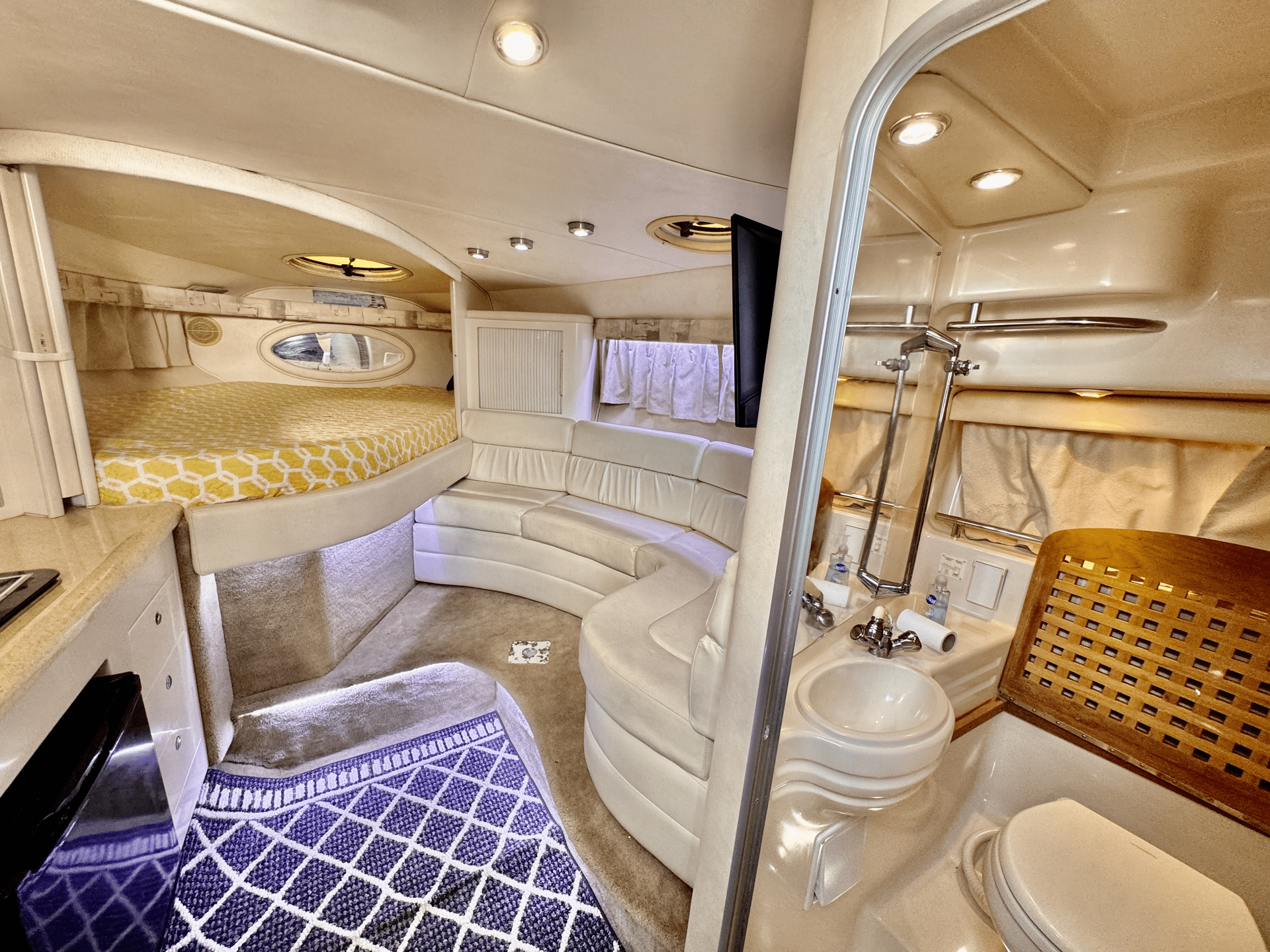 Interior of a yacht cabin with a bed, seating area, and bathroom. Mostly white and beige colors.