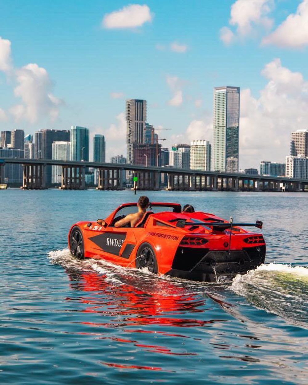 Red car-boat cruising on water near a city skyline. Man driving. Bright blue water and sky.