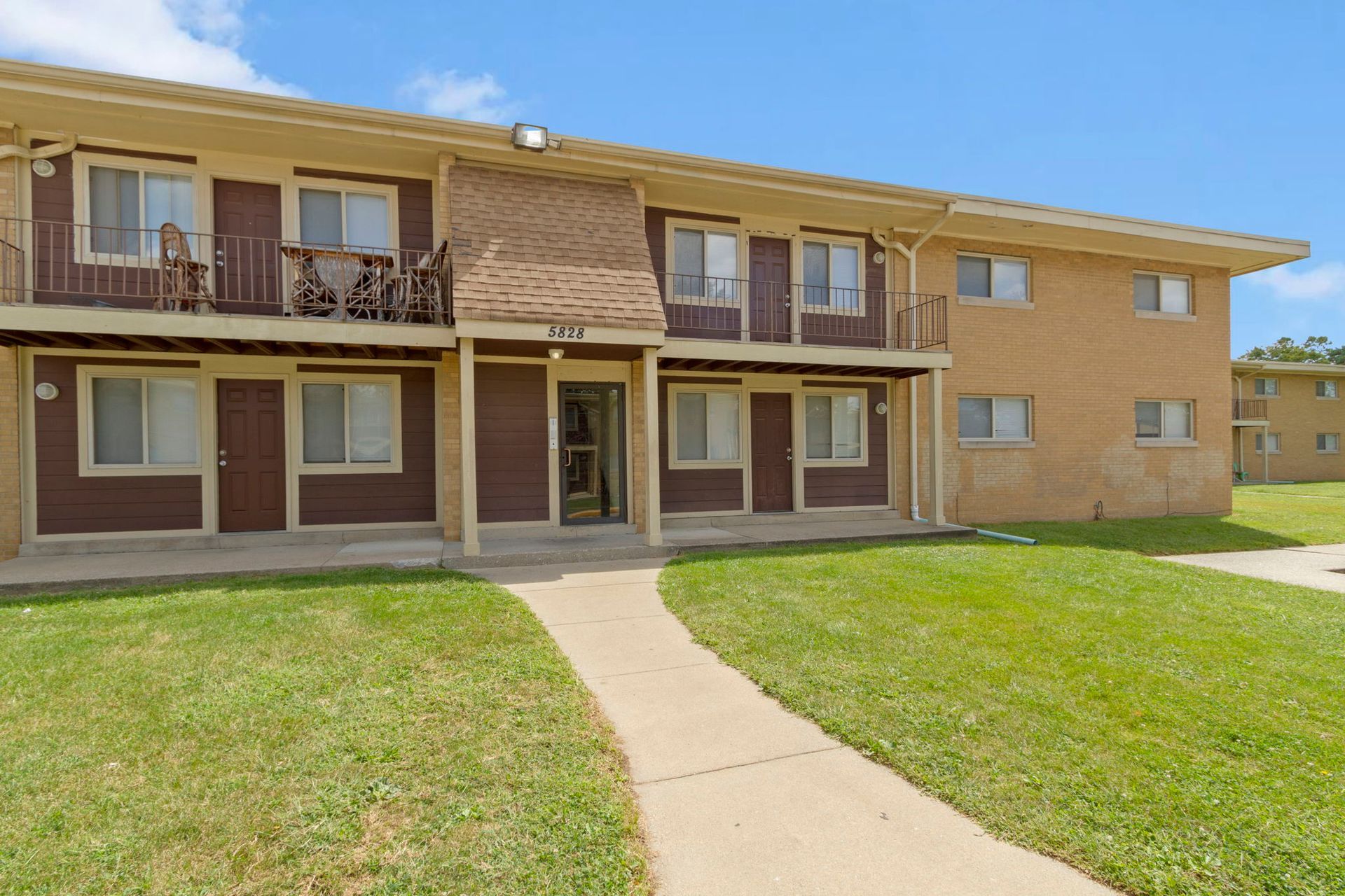 Apartment building with brown trim and balconies. Green grass and a sidewalk lead to the entrance on a sunny day.