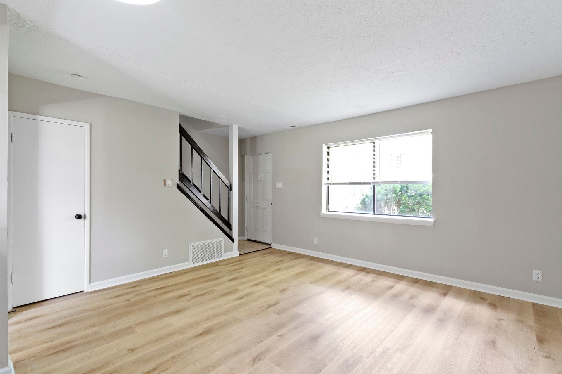 Empty living room with light wood floors, stairs, and window.