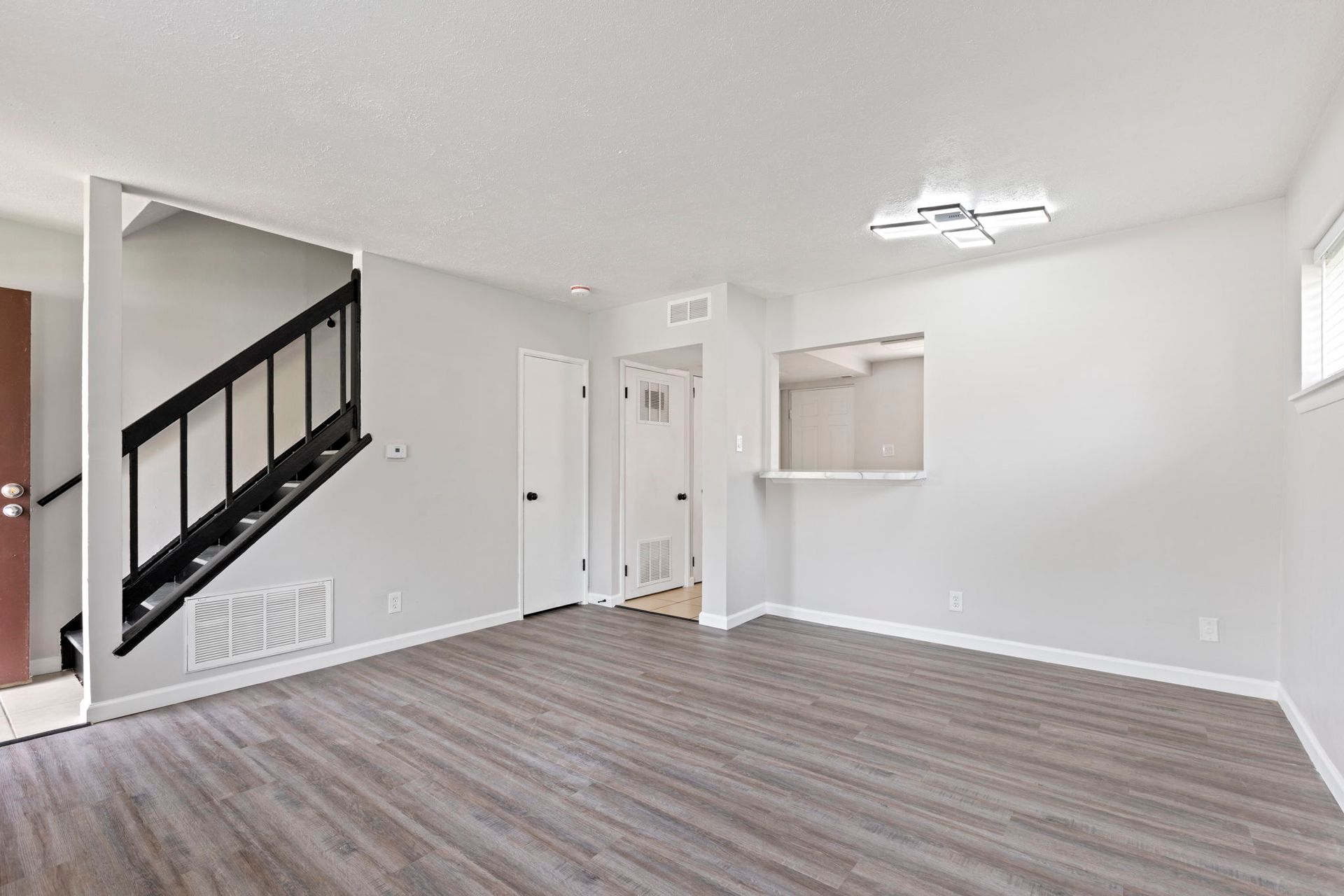 Empty living room with gray wood flooring, white walls, stairs, and an open kitchen bar.