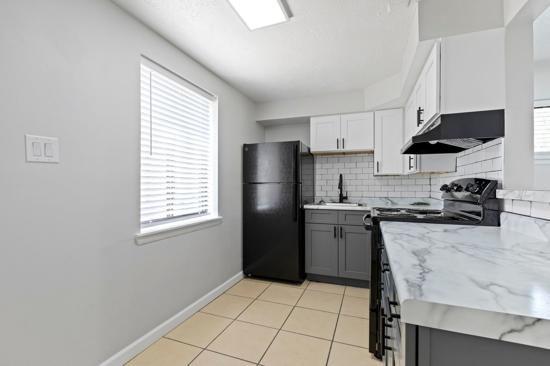 A kitchen with white and gray cabinets, black appliances, and marble countertops; window with blinds.