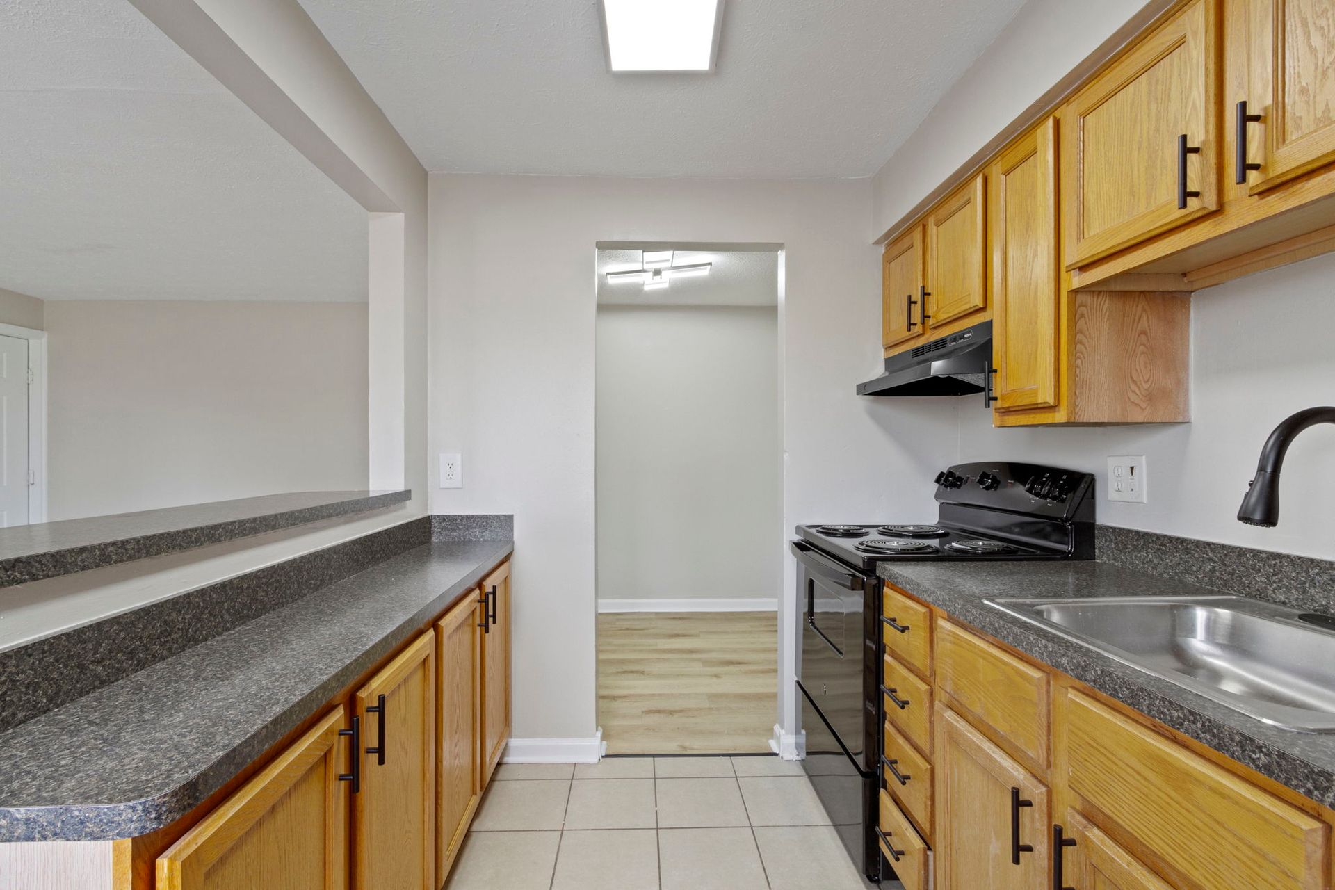 Kitchen with wooden cabinets, black appliances, and grey countertops. View leads to a hallway and living space.