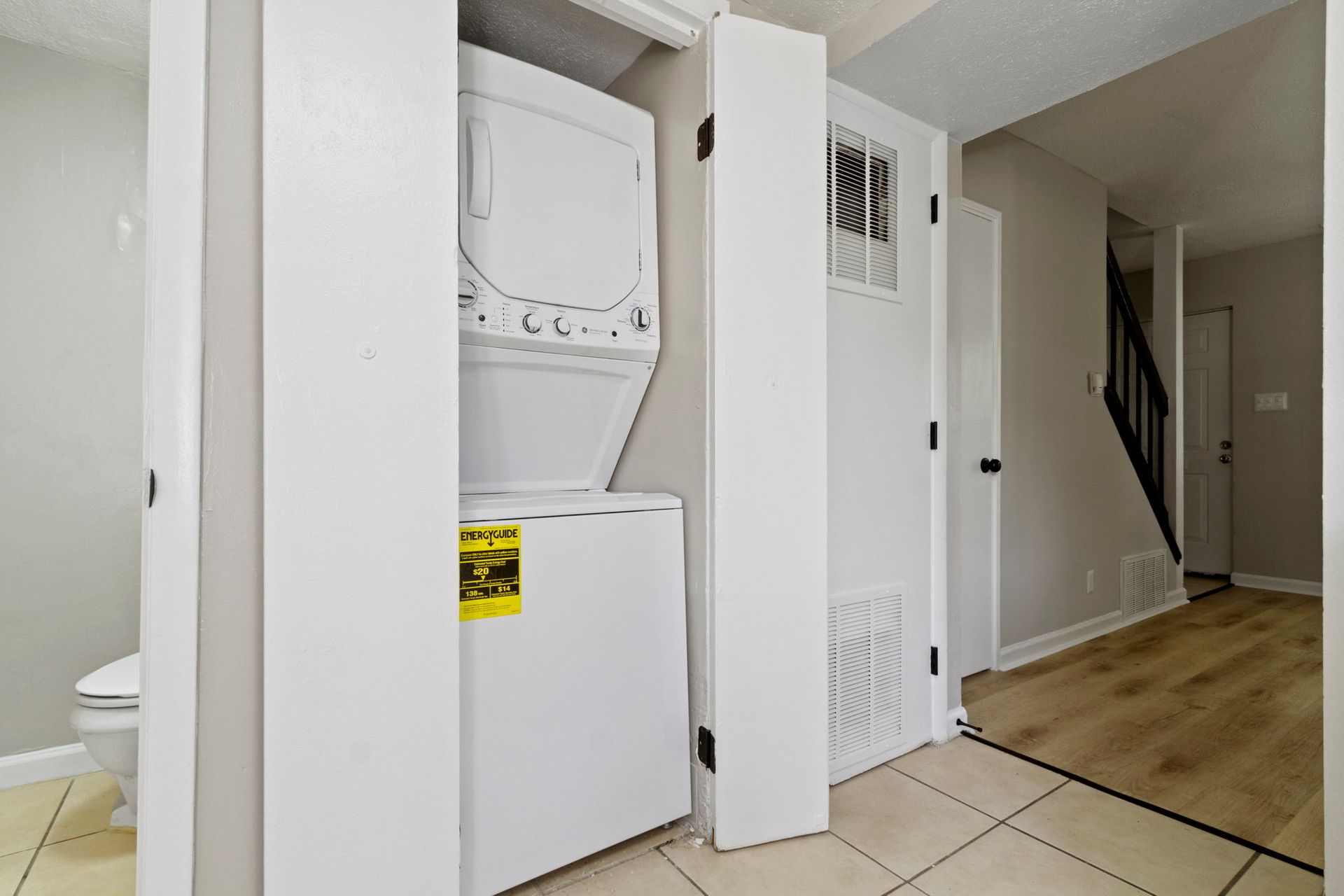 Stacked washer and dryer in a white closet, next to an air vent and stairs.