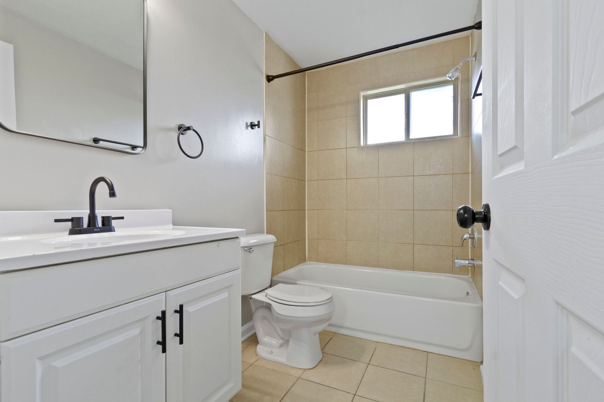 Bathroom with white vanity, toilet, bathtub, and light beige tile. Black fixtures.