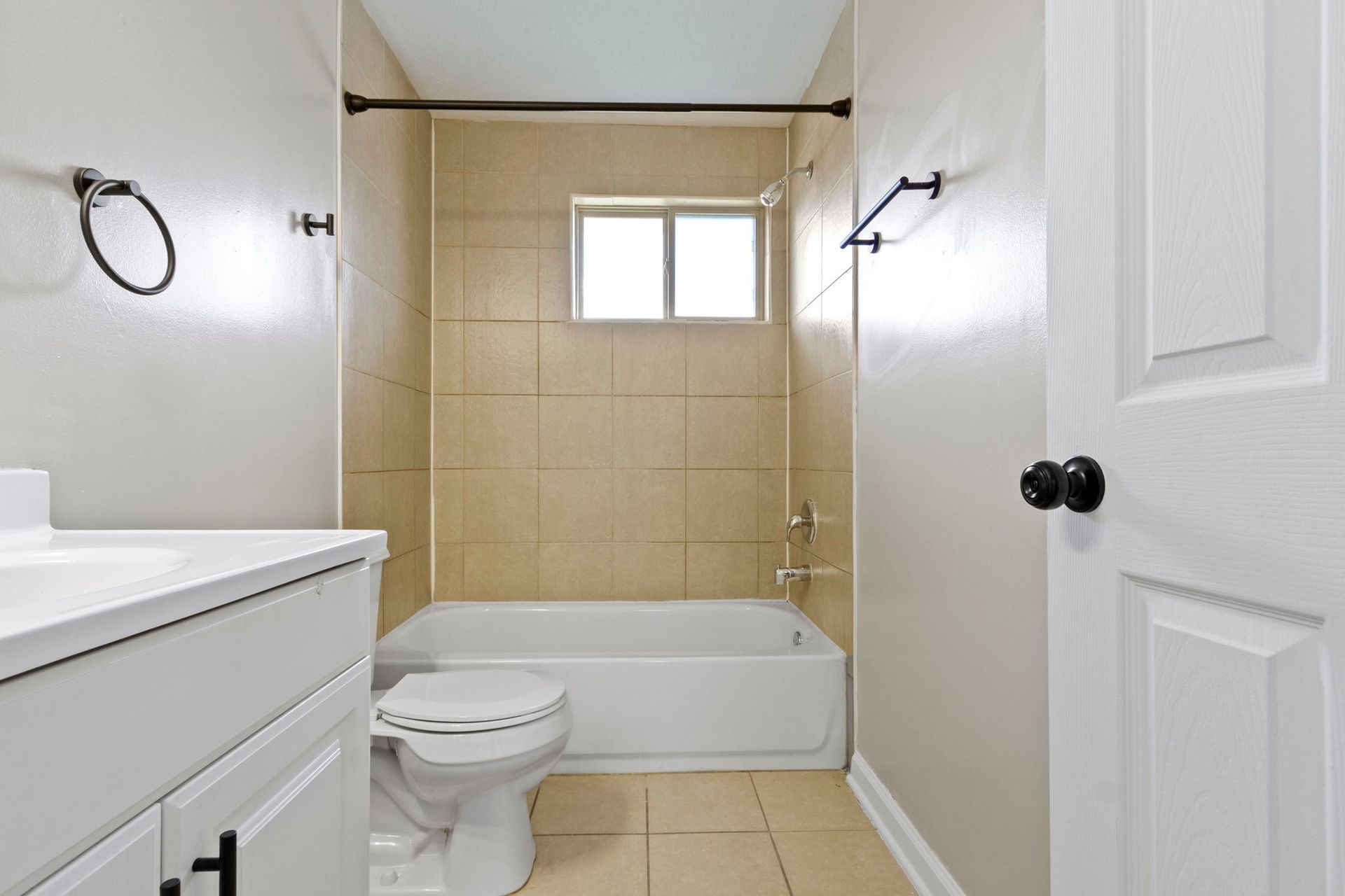 Bathroom with a white tub, toilet, and vanity, and beige tile.  Black fixtures on gray walls.