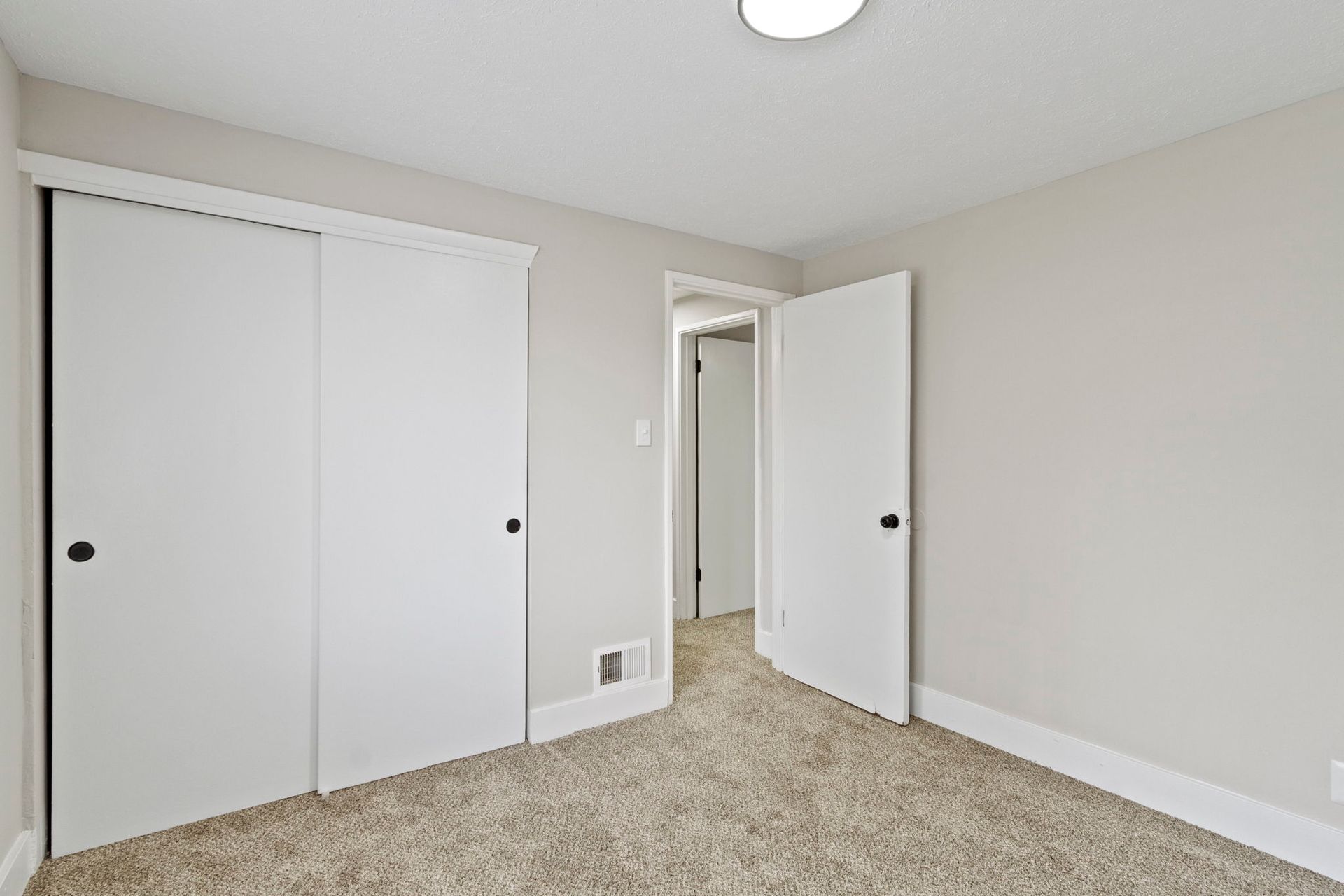 Bedroom interior with white closet doors and a partially open door to a hallway. Light tan carpet.