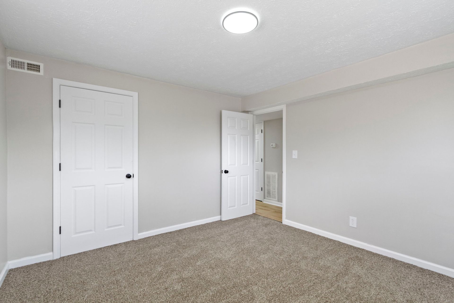 Empty bedroom with light gray walls, white doors, beige carpet, and overhead lighting.