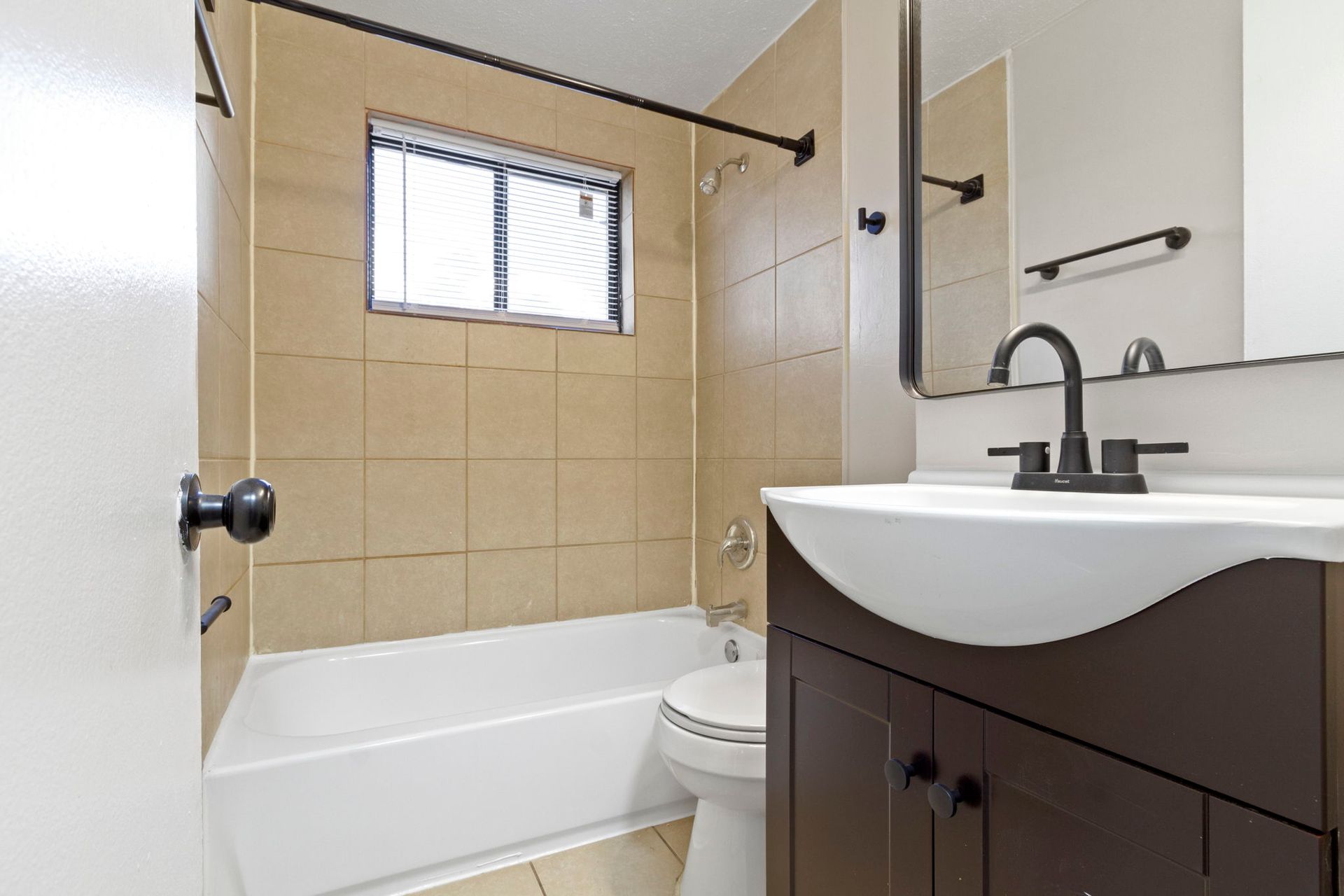 Bathroom with brown vanity, beige tile, white tub, and small window.
