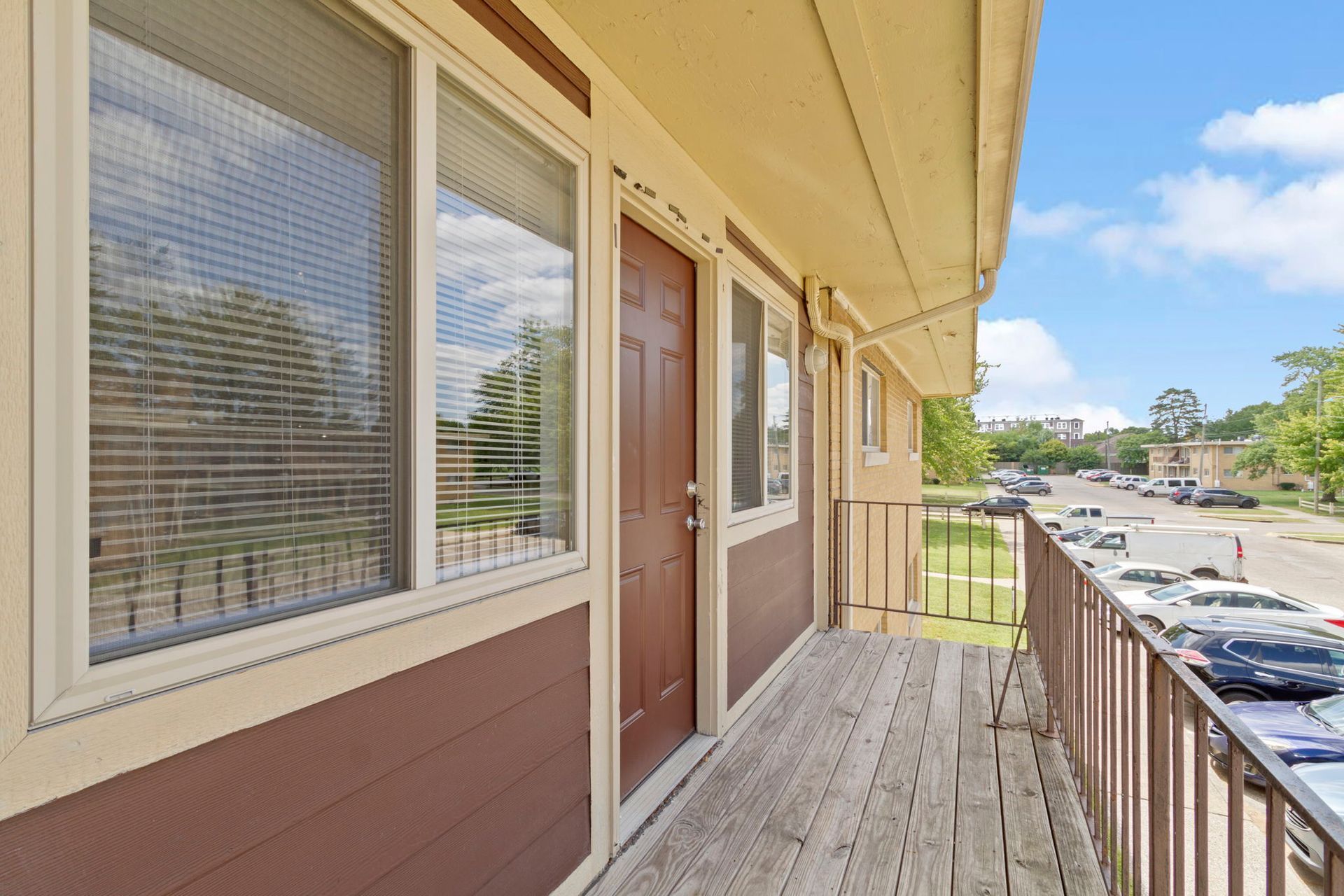 Balcony exterior of an apartment with brown door, white-framed windows, wooden floor, and a street view.