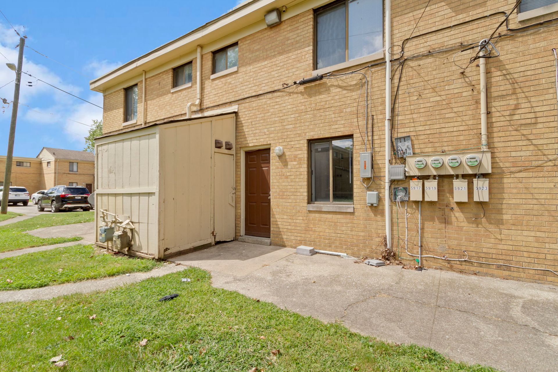Back exterior of a tan brick apartment building with a small wooden storage shed and utility boxes.