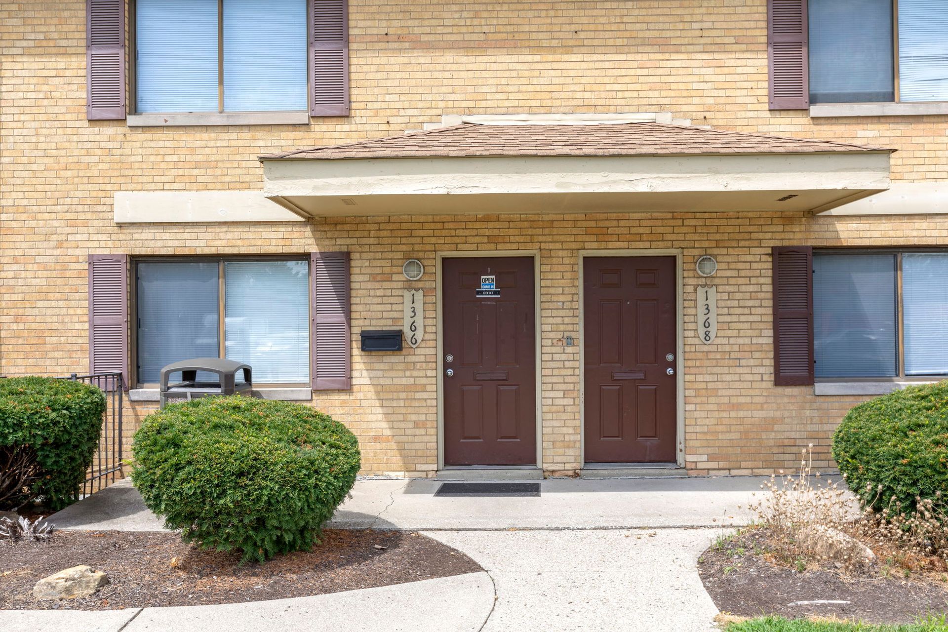 Tan brick apartment building with two dark brown doors, shutters, and shrubs.