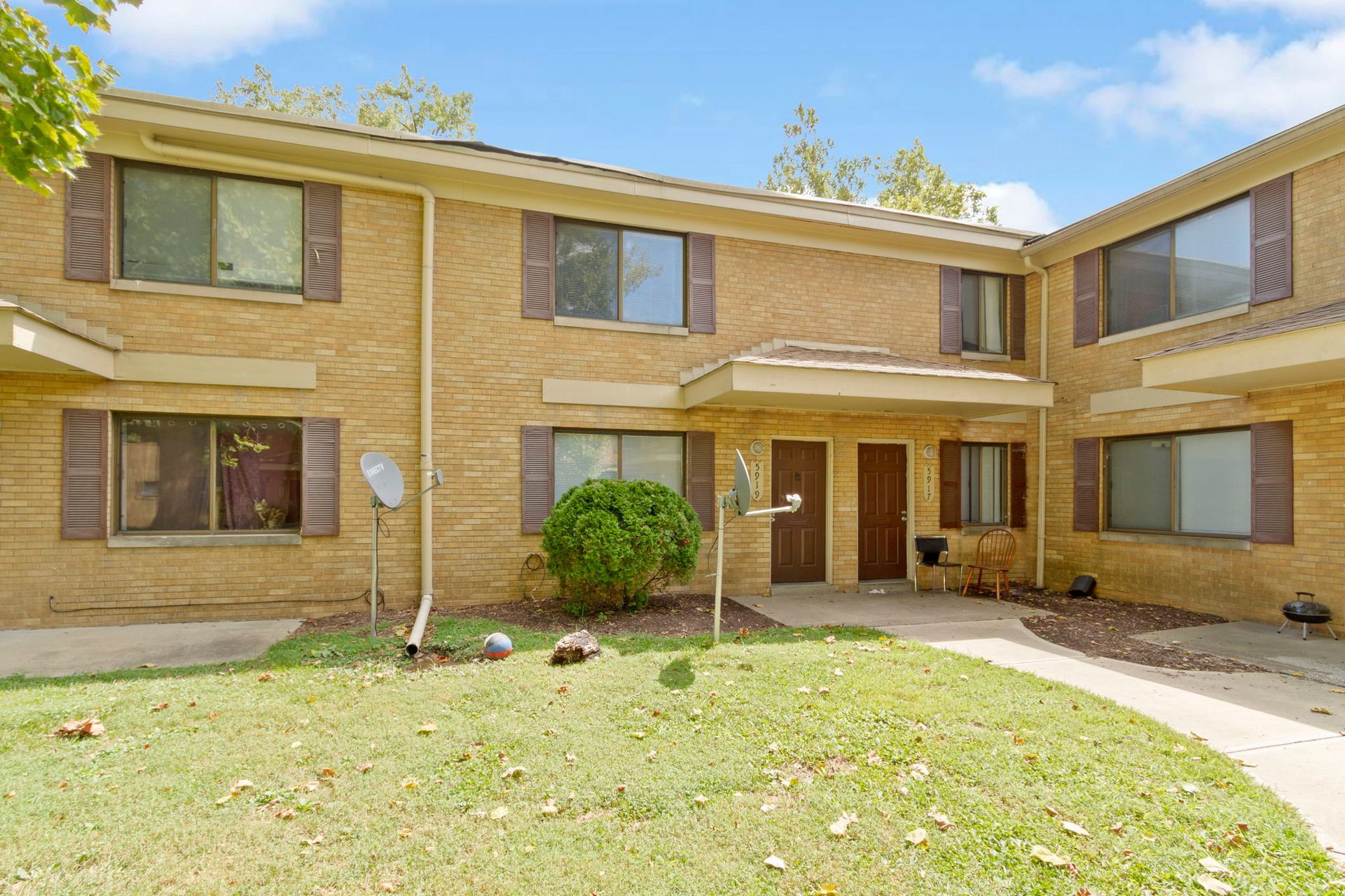 Two-story brick townhouses with brown shutters and doors, a small yard, and a blue sky.