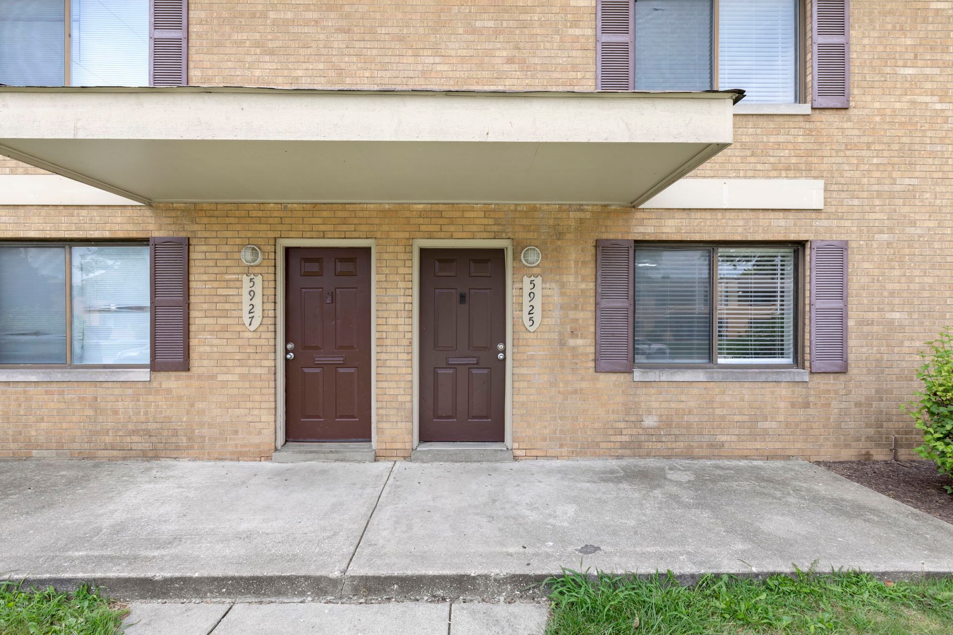 Apartment building exterior: brown doors, beige brick, windows with shutters, concrete walkway.