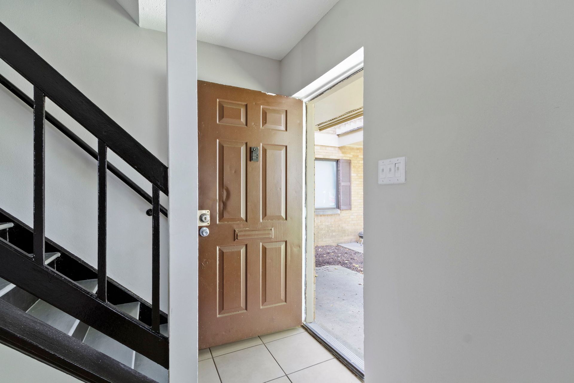Open brown door in gray-walled entryway, stairs to the left, leading outside.