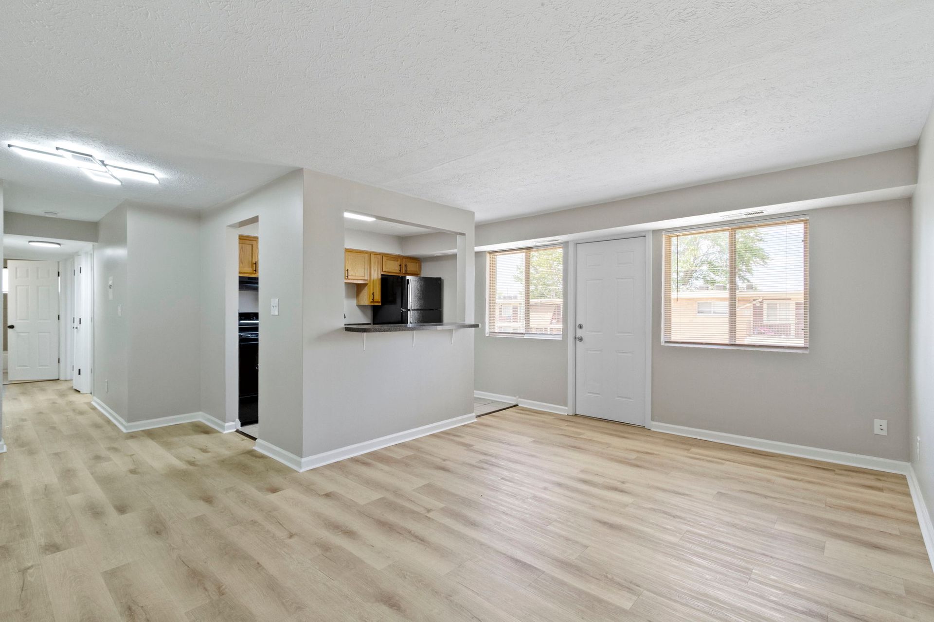 Spacious living room with light wood floors, kitchen pass-through, and large windows letting in sunlight.