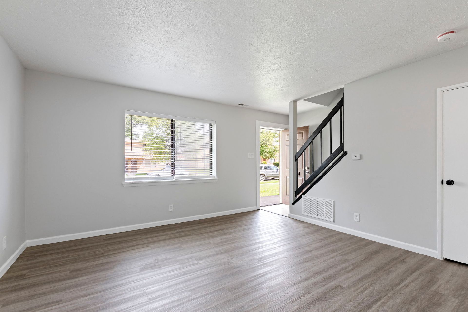 Empty living room with gray walls, wood-look flooring, window, open doorway to exterior, stairs.