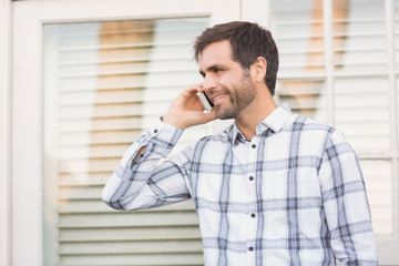 Man in plaid shirt talking on a cell phone, smiling, near a window with white shutters.