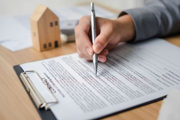 Person signing a real estate contract with a pen; model house in the background.