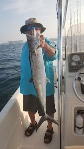 Man on a boat holding up a large, silver king mackerel he caught in the ocean.