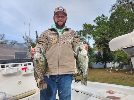 Man on a boat holding two crappie fish; wearing a hat and jacket, smiling. Green trees in the background.