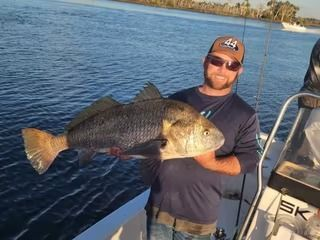 Man on boat holds large black drum fish, blue water background.