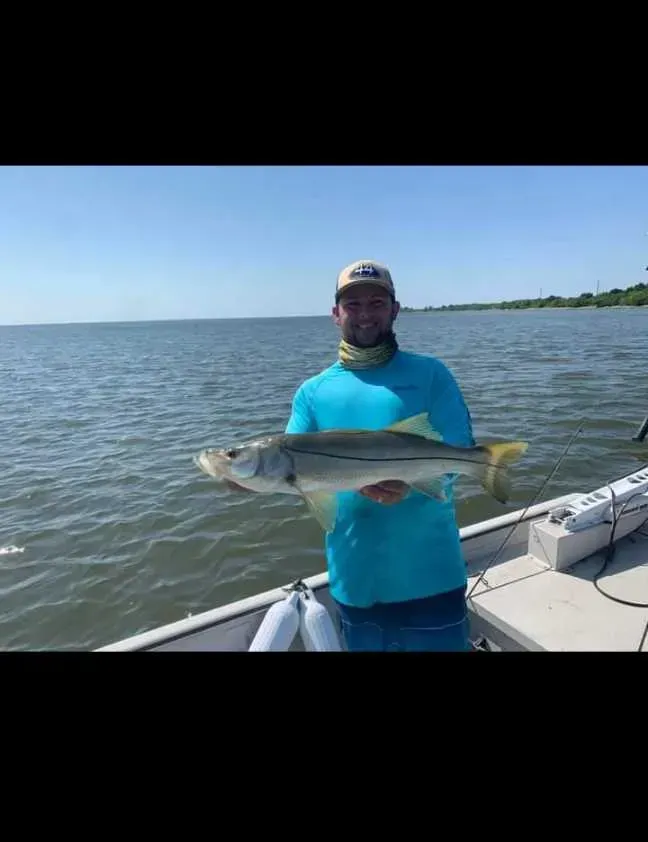 Man on a boat holding a snook fish, blue water, clear sky.