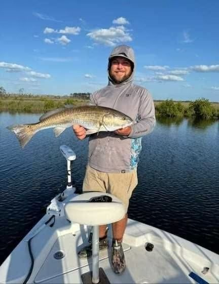 Man on boat holds up a redfish he caught, smiling in front of a marsh with blue sky.