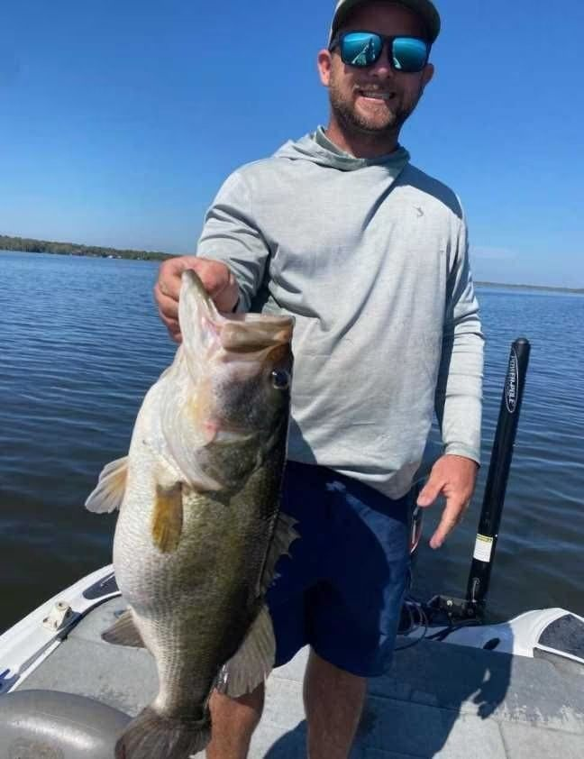 Man in sunglasses holding a large largemouth bass on a boat, smiling; water and blue sky background.