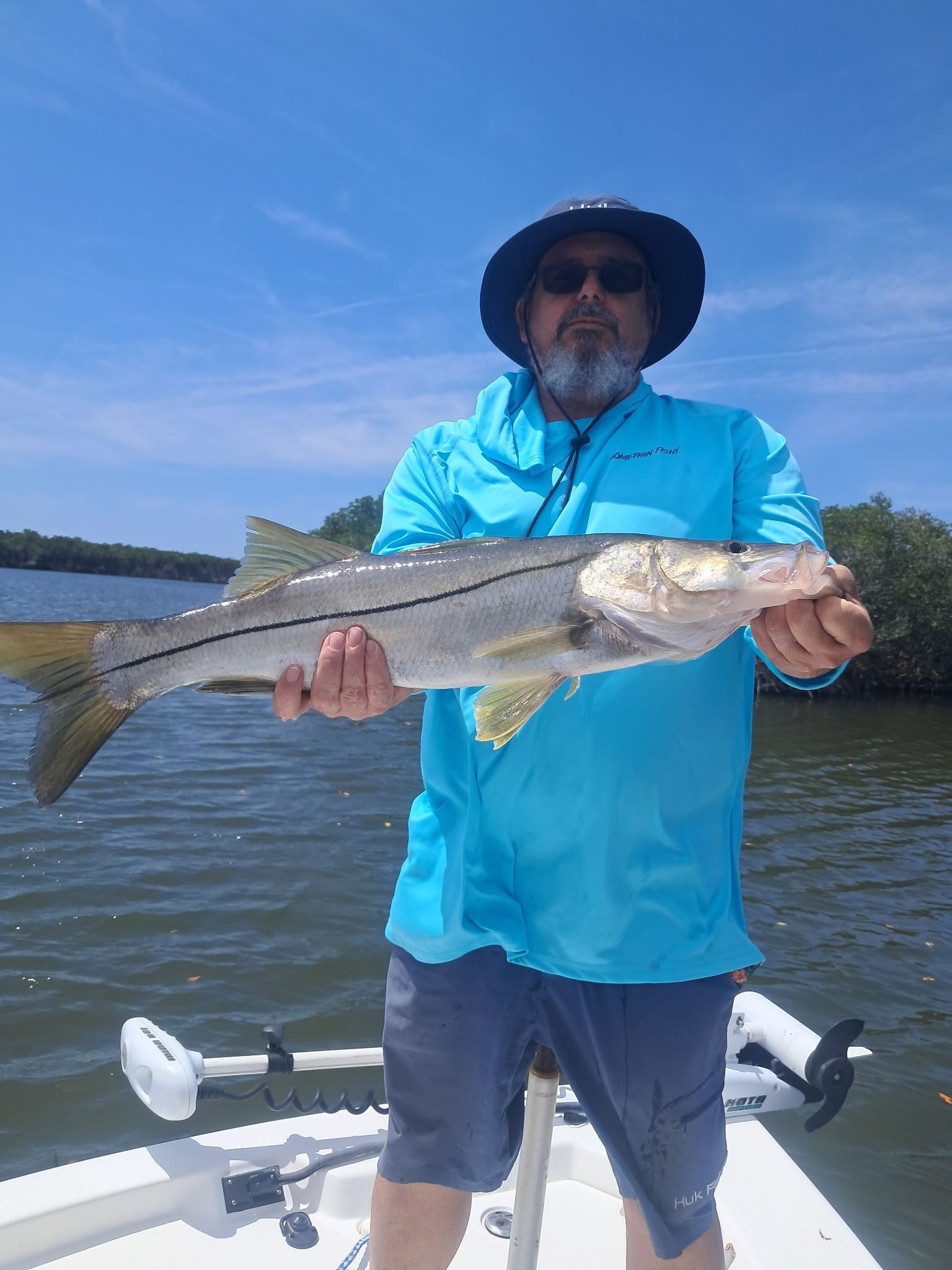 Man on a boat holds a large, reddish-silver fish with an open mouth, against a blue sky and water backdrop.