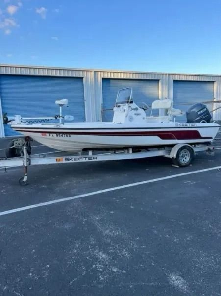 White and maroon fishing boat on a trailer in front of storage units under a blue sky.
