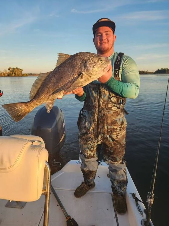 Man on boat holds large black fish he caught. Wearing fishing gear, smiles at the camera.