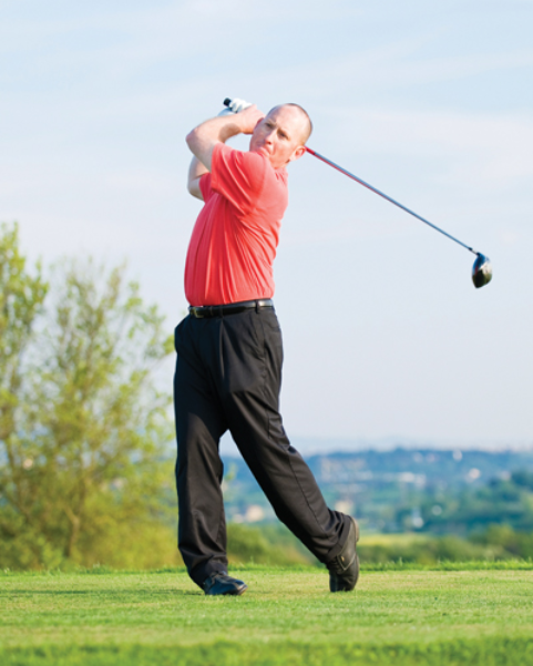 Golfer in red shirt and black pants finishing a golf swing on a grassy course