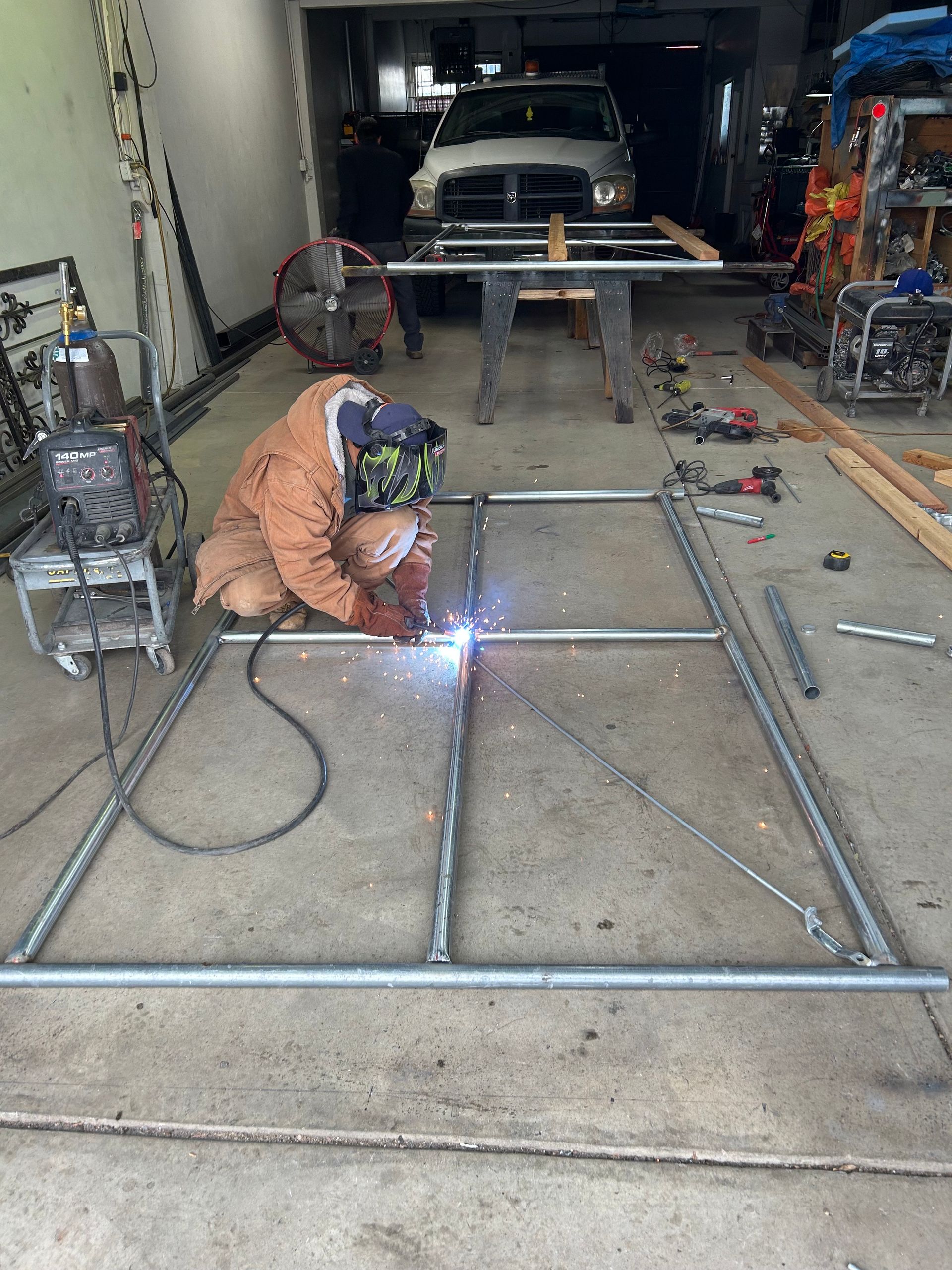 A person welding metal frame in a garage. They wear protective gear. Tools and a truck are in the background.