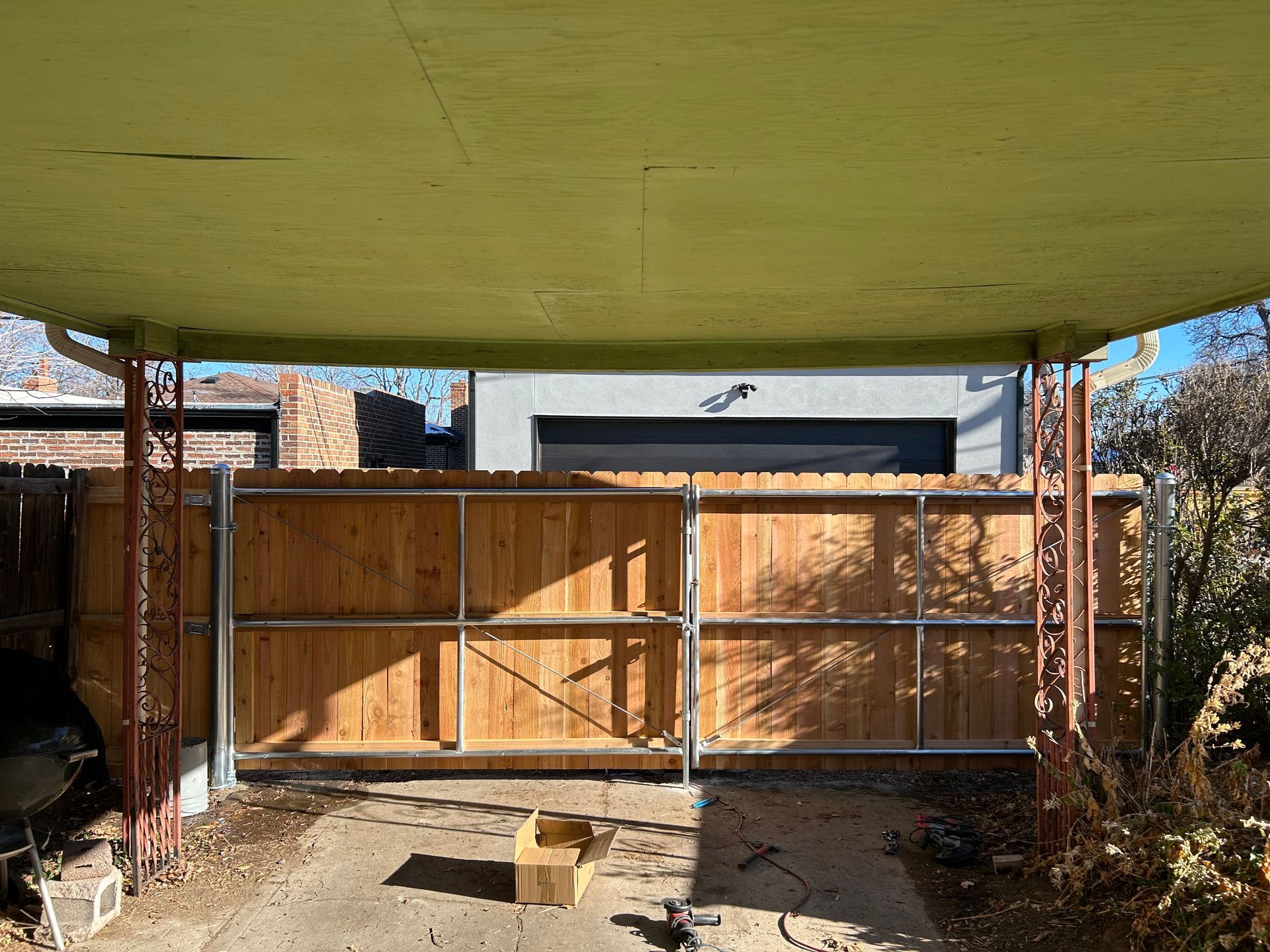 A wooden fence and gate under a yellow-painted carport, with a box and tools on the ground.