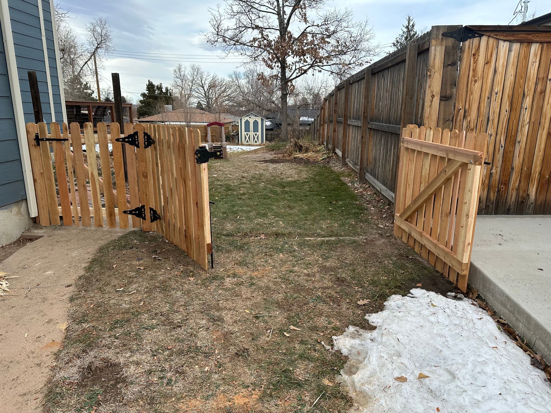 Wooden gates leading into a backyard with patchy grass and snow. Wooden fence on the right side.