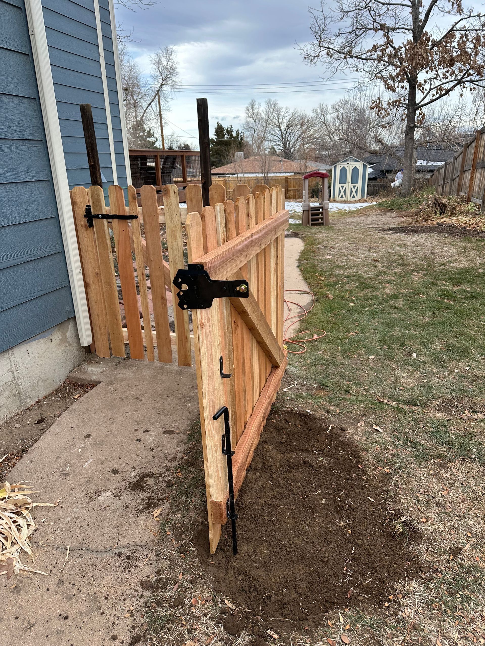 Wooden gate in a backyard, partially open. Brown fence with black hardware near a house.