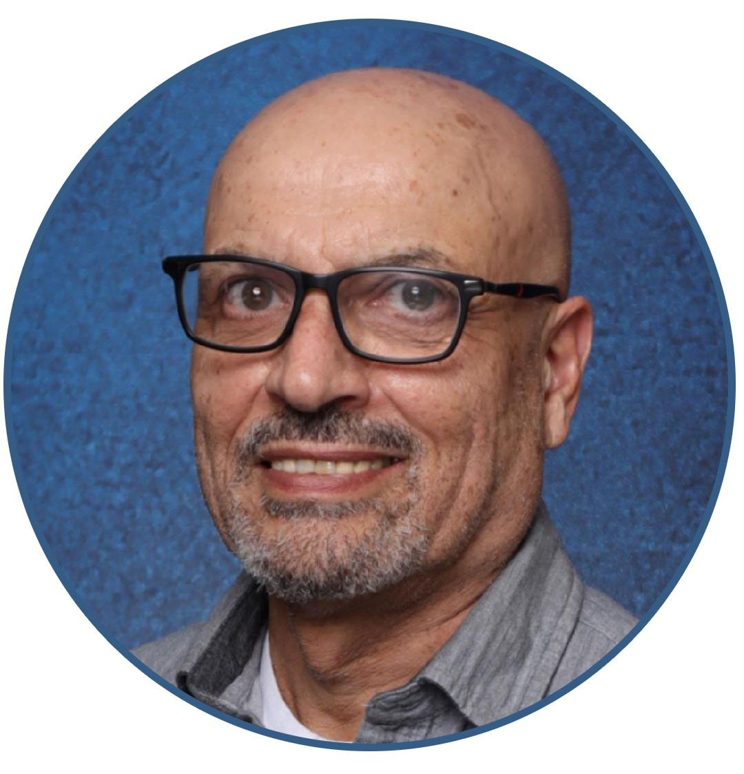 Bald man with glasses smiling, wearing a grey shirt, in front of a blue textured background.