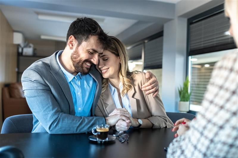 Smiling couple embraces across table from person in office setting.