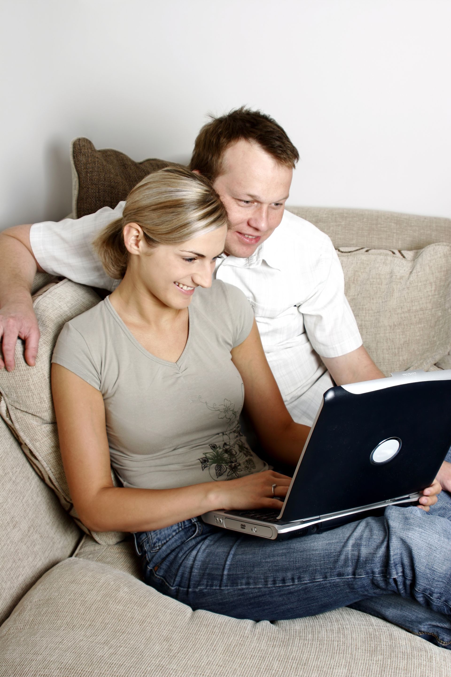 Couple on a sofa looking at a laptop, woman typing, man smiling, indoor setting.