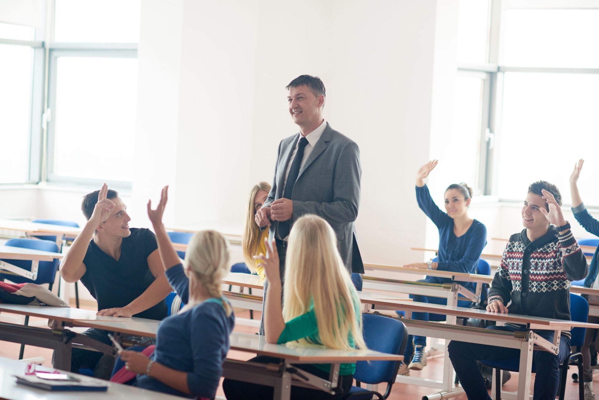 A man is standing in front of a classroom full of students raising their hands to answer a question.