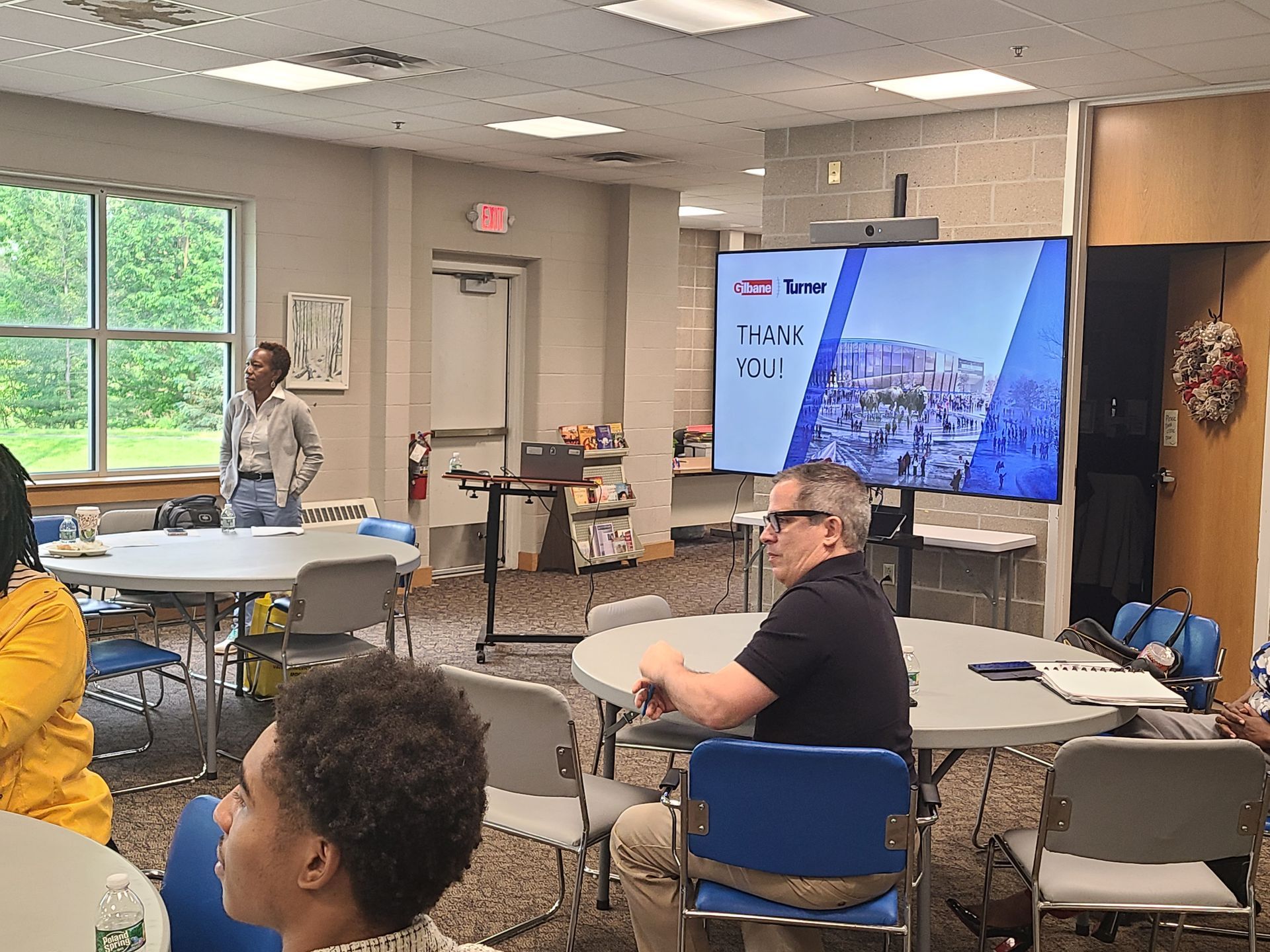 A group of people are sitting at tables in a room with a large screen on the wall.